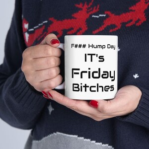 May include: A white ceramic mug with the text "F### Hump Day, IT's Friday Bitches" in black. The mug is held by a person wearing a dark blue sweater with a red reindeer pattern.