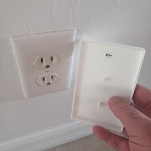 May include: A white electrical outlet with two sockets is mounted on a white wall. A white outlet cover is held nearby, with the word "TOP" printed on it. The cover has two small pegs.