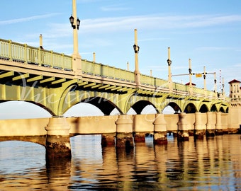 May include: A scenic photograph of a bridge over water. The bridge has arched supports and is painted in shades of green and beige. The water reflects the bridge and the sky, creating a symmetrical image.