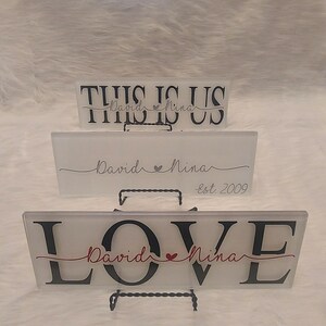 May include: Three frosted glass signs with black lettering and a red heart. The top sign says "This is Us" with the names "David" and "Nina" underneath. The middle sign says "David Nina" with a heart and "Est 2009" underneath. The bottom sign says "LOVE" with the names "David" and "Nina" underneath.