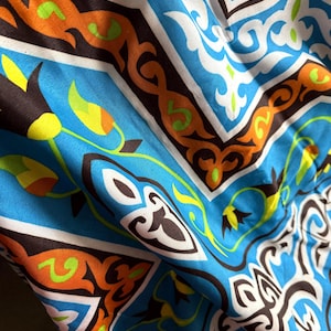 Puede incluir: Primer plano de una tela con un patrón vibrante e intrincado. El diseño presenta un fondo azul brillante con formas geométricas y elementos florales en blanco, naranja, marrón y verde. La tela parece suave y fluida.