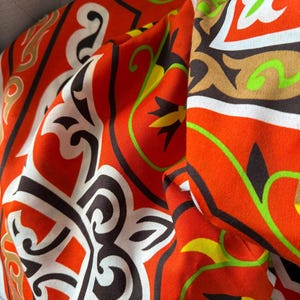 Könnte beinhalten: Nahaufnahme eines leuchtend orangefarbenen Stoffes mit einem sich wiederholenden Muster aus weißen, braunen und schwarzen geometrischen Formen und floralen Akzenten. Das Design enthält grüne und gelbe Details und schafft einen auffälligen Textil.