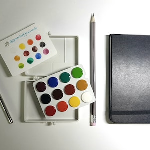Puede incluir: Una paleta de acuarela de plástico blanco con 12 pocillos redondos, un cuaderno de bocetos negro, un lápiz y un pincel. La paleta está abierta y los pocillos están llenos de diferentes colores de pintura de acuarela. El cuaderno de bocetos está cerrado y tiene una cubierta negra. El lápiz está afilado y tiene una goma de borrar rosa. El pincel es plateado y tiene un mango negro.