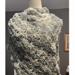 Può includere: Uno scialle fatto a mano, a trama aperta, nelle tonalità del grigio e del crema, esposto su un manichino. Lo scialle presenta un design traforato e strutturato, perfetto per aggiungere un tocco di eleganza a qualsiasi outfit. I colori tenui e neutri lo rendono un accessorio versatile.
