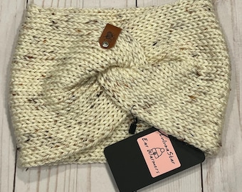 May include: Cream-colored knitted ear warmer with a twisted design. The ear warmer has flecks of brown and black throughout the yarn. A small brown leather tag with a silver button is attached. A black tag with the text "Luluna Star Ear Warmers" is also attached.