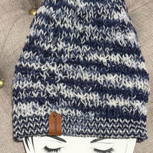 May include: A knitted beanie hat in navy, white, and light blue. The hat has a textured knit design and a small brown leather tag with the text "lulunastar". The beanie is displayed on a white card with drawn eyelashes.