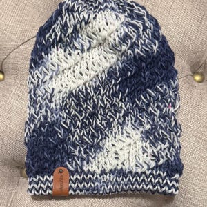 May include: A knitted beanie hat in navy blue, white, and pink. The hat has a textured stitch pattern and a small brown leather tag with the text "lulunaStar". This hat is designed for warmth and style, perfect for colder weather.