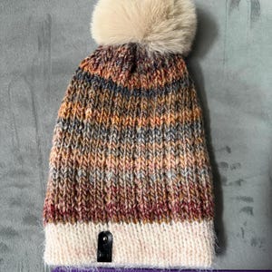 May include: A hand-knitted beanie hat with a cream-coloured faux fur pom-pom. The hat features a colour gradient of brown, orange, and grey. The bottom band is cream-coloured. A black tag is sewn on the bottom. The hat is approximately 25 cm long.