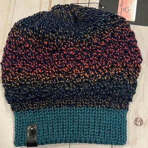 Könnte beinhalten: Eine handgestrickte Beanie-Mütze mit einem Farbverlauf von Marineblau zu Orange und Rosa, der in einen türkisfarbenen Rippenbund übergeht. Ein kleines schwarzes Etikett mit Logo ist am Bund befestigt. Die Mütze ist aus weichem Garn.