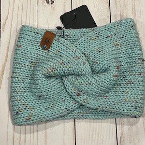 May include: A light blue knitted headband with a twisted design. The headband has flecks of color throughout and a small brown leather tag with a button. The tag has a small design on it.