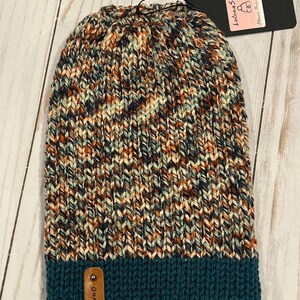 Könnte beinhalten: Eine handgestrickte Mütze in einer Mischung aus Farben wie Orange, Türkis, Marineblau und Creme. Das untere Band ist türkis, und ein kleines Lederetikett trägt die Aufschrift "MADE". Ein schwarzes Etikett trägt die Aufschrift "Luluna Star Messy Bun Beanie".