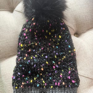 May include: A black knitted beanie with colourful flecks of pink, yellow, and blue. The hat has a grey ribbed brim and a large black faux fur pom-pom on top. A small black tag is attached to the brim.