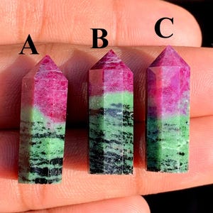 Può includere: Tre punte di cristallo di rubino in zoisite lucidate. Ogni cristallo presenta una parte superiore rosa, una sezione centrale verde e una base nera e verde. I cristalli sono tenuti in una mano.