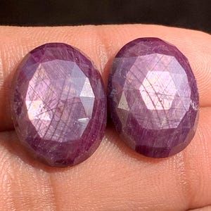 May include: Two oval-cut, deep reddish-purple gemstones. The gems are held in a hand, and the facets reflect light, creating a sparkling effect. The gems appear to be rubies.