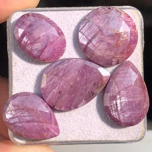 Può includere: Cinque gemme rosa sfaccettate, ovali e a goccia, sono esposte in un piccolo contenitore quadrato bianco. Le gemme hanno un aspetto traslucido con venature interne. Le gemme sono tenute dalle dita di una persona.
