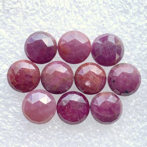 May include: Ten round, faceted gemstones in shades of red and pink. The gems have a polished surface and are arranged together. These are likely rubies, suitable for jewellery making. Each gem is approximately 1 cm in diameter.