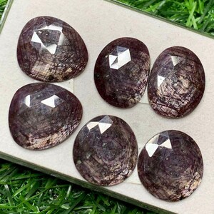 Peut inclure: Six pierres précieuses facettées de forme ovale, de couleur violet foncé et avec un reflet argenté. Les pierres sont disposées en cercle sur une surface blanche.
