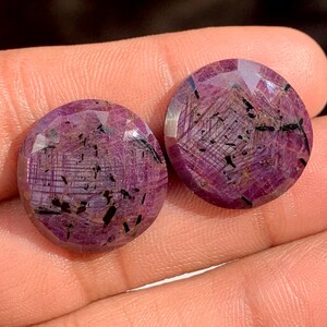 May include: Two round, faceted gemstones with a deep magenta hue. The stones have black inclusions and a network of fine lines within. The gems are held in a hand, showcasing their polished surfaces and intricate internal patterns.