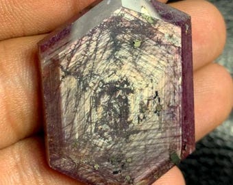Puede incluir: Una gema hexagonal facetada de color púrpura intenso. La piedra presenta patrones internos de líneas oscuras e inclusiones, con matices más claros. Se ven pequeñas motas de material dorado.