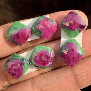 May include: Six faceted gemstones with a square shape. The gems have a vibrant pink center surrounded by a green border. The gems are displayed on white paper.