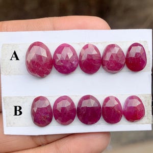 Puede incluir: Una colección de rubíes facetados de forma ovalada, dispuestos en dos filas etiquetadas A y B. Los rubíes muestran un tono rojo intenso y están colocados sobre un fondo blanco. Las piedras preciosas se muestran en un primer plano.