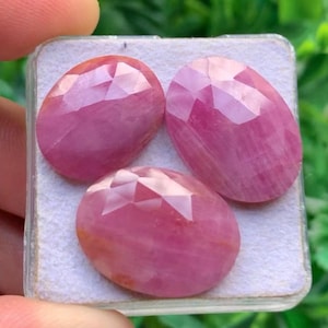 Può includere: Tre pietre preziose rosa di forma ovale e sfaccettate, disposte su un cuscinetto espositivo bianco. Ogni pietra presenta una superficie lucida con sfaccettature geometriche che riflettono la luce, mostrando una tonalità rosa vibrante.
