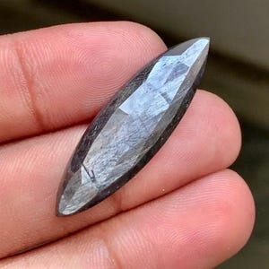 May include: A marquise-cut gemstone with a dark grey, metallic, and charcoal-coloured surface. The stone features a faceted finish that creates a reflective, textured appearance with visible internal inclusions and a sharp, pointed shape.