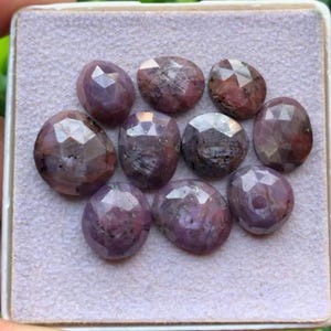 May include: A collection of nine oval-shaped, faceted rubies displaying deep shades of purple, pink, and red. Each gemstone features a polished, multi-faceted surface that reflects light, arranged in a cluster on a light grey, textured display pad.