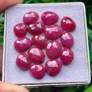 Pode incluir: Uma coleção de treze rubis facetados de formato oval com um tom vermelho rosado profundo. Cada pedra preciosa apresenta uma superfície polida com facetas geométricas que refletem a luz, dispostas sobre uma almofada de exibição cinza claro texturizada.