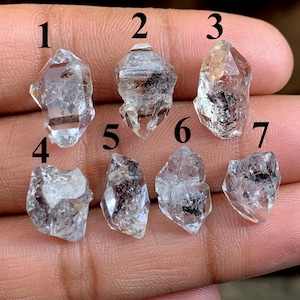 Peut inclure: Sept pointes de cristal de quartz clair de formes et de tailles variées sont présentées. Les cristaux présentent des inclusions et leur couleur varie du transparent au brun clair. Les cristaux sont tenus dans une main, mettant en valeur leur beauté naturelle et leurs formations uniques.
