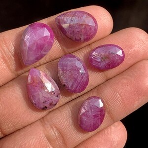 Peut inclure: Six pierres précieuses rubis facettées de différentes formes, dont ovales et en goutte. Les gemmes sont d'un rouge-rosé profond, avec des structures internes visibles et des reflets de lumière. Les pierres sont présentées sur une surface claire.