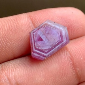 Peut inclure: Une pierre précieuse facettée en forme de bouclier avec une teinte rose et violette translucide. La pierre présente une taille géométrique à plusieurs niveaux avec des lignes internes distinctes et une surface polie, montrant un mélange de zones de couleur violet clair et foncé.