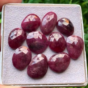 May include: A collection of ten oval-shaped, faceted rubies with a deep red color. Each gemstone features a polished surface with geometric cuts that reflect light, arranged together on a light gray, textured display pad within a square container.