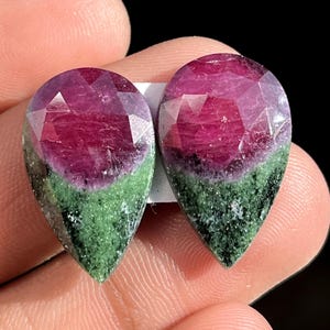 Puede incluir: Dos gemas en forma de pera con la parte superior facetada. Las gemas presentan un gradiente de color vibrante, que va del magenta intenso en la parte superior al verde moteado en la base. Las gemas están sobre un fondo oscuro.