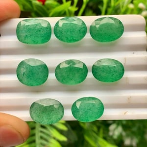May include: Eight oval-shaped green gemstones with faceted surfaces arranged in three rows on a white display tray. Each stone features a translucent appearance with subtle internal speckling and a polished finish reflecting light.