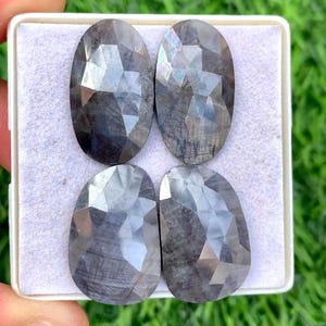 May include: Four faceted, oval-shaped gemstones with a dark gray color and a polished finish. The gems are arranged in a white tray against a backdrop of green grass. The gemstones have a reflective surface.