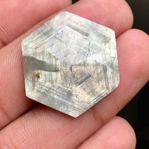 May include: A hexagonal, translucent gemstone with a pale gray and white color palette. The surface features visible internal inclusions, linear striations, and a faceted top that creates a geometric pattern across the stone's flat, six-sided shape.