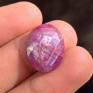 May include: A faceted, oval ruby gemstone. The gem has a deep pinkish-red hue with internal inclusions and a polished, reflective surface. The cut reveals multiple facets, enhancing its brilliance and colour.