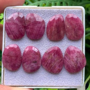 Pode incluir: Oito rubis rosa facetados em forma de lágrima, dispostos em duas filas de quatro dentro de uma caixa branca. Cada pedra preciosa apresenta uma superfície polida com inclusões naturais visíveis e uma cor magenta rosada intensa e vibrante.