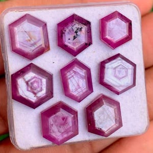 Pode incluir: Uma coleção de sete pedras preciosas hexagonais facetadas em tons de rosa e magenta. Cada gema, provavelmente rubis, exibe padrões internos únicos. Perfeitas para a criação de joias.