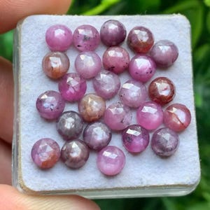Puede incluir: Una colección de veintidós piedras preciosas redondas y facetadas de color rosa y púrpura rojizo, dispuestas en una cuadrícula sobre una base de espuma blanca. Cada piedra presenta una superficie multifacetada que refleja la luz.