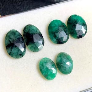 May include: Six oval-shaped emerald gemstones with faceted surfaces. The gems display a deep green color with dark inclusions, and are arranged on a light-colored surface. The stones vary slightly in size and shape.