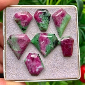 Può includere: Una collezione di sette gemme sfaccettate di rubino e zoisite disposte su un supporto espositivo grigio chiaro. Ogni pietra presenta una combinazione unica di inclusioni minerali in rosa magenta vibrante e verde bosco profondo.