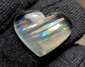 Pode incluir: Uma pedra preciosa iridescente em forma de coração com raios de arco-íris. A pedra transparente tem uma superfície polida, refletindo a luz com cores azul, amarelo e rosa. A pedra preciosa é segurada contra um fundo escuro e texturizado.