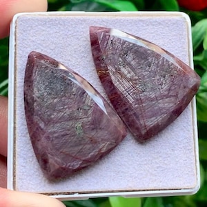Può includere: Due pietre preziose triangolari sfaccettate con una tonalità rosso-viola intenso e inclusioni interne visibili a forma di ago. Le pietre sono lucidate con una superficie liscia e sono esposte fianco a fianco su un cuscinetto in schiuma grigio chiaro.