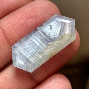 Pode incluir: Um cristal hexagonal alongado e translúcido com um tom azul pálido e inclusões fibrosas brancas no interior. A pedra preciosa apresenta bordas facetadas e uma superfície polida, exibindo uma estrutura geométrica clara com padrões internos.