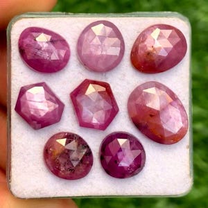 Puede incluir: Colección de siete gemas facetadas, de forma ovalada y geométrica, en tonos rosas y rojos. Las gemas se muestran en un recipiente blanco, con un fondo verde.
