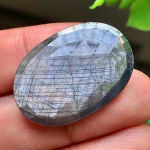 Peut inclure: Une pierre précieuse à facettes de forme ovale avec une teinte bleu gris translucide. La surface présente un motif distinct d'inclusions linéaires sombres et entrecroisées qui créent un aspect de grille sur le dessus poli et facetté.