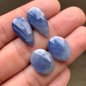 Può includere: Quattro pietre preziose di zaffiro blu sfaccettate in varie forme: due a goccia e due ovali. Le gemme sono tenute in una mano, mostrando le loro superfici lucide e le variazioni di colore. Gli zaffiri hanno un aspetto traslucido.