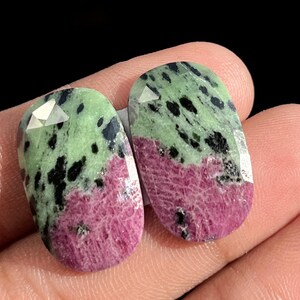 Può includere: Due pietre preziose di zoisite di forma ovale. La metà superiore di ogni pietra è verde chiaro con macchie nere e la metà inferiore è rosa intenso. Le pietre hanno un bordo superiore sfaccettato.